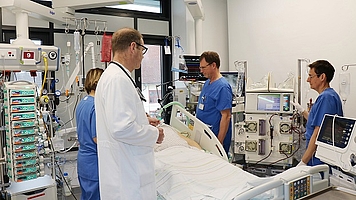 Bett auf Intensivstation mit viel Medizintechnik umgeben. ein Mann im weißen Kittel und 3 Pflegekräfte in blauer Bekleidung kümmen sich um patient. 